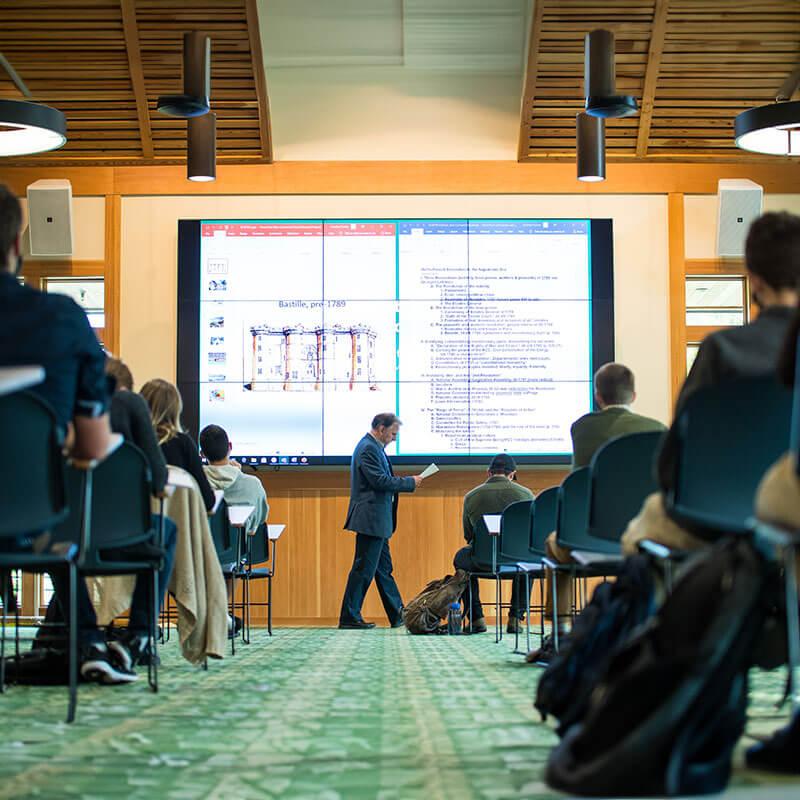 a professor walks in front of a screen surrounded by a small group of students at desks