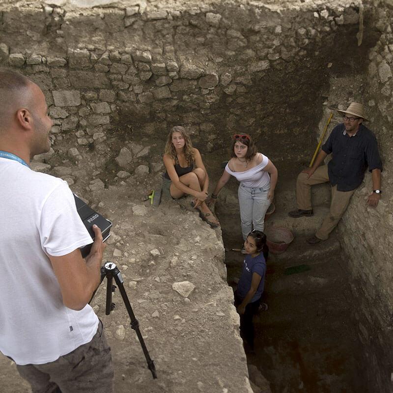 three people stand in a stone stairwell on an archeological dig site, a third person sets up a tripod