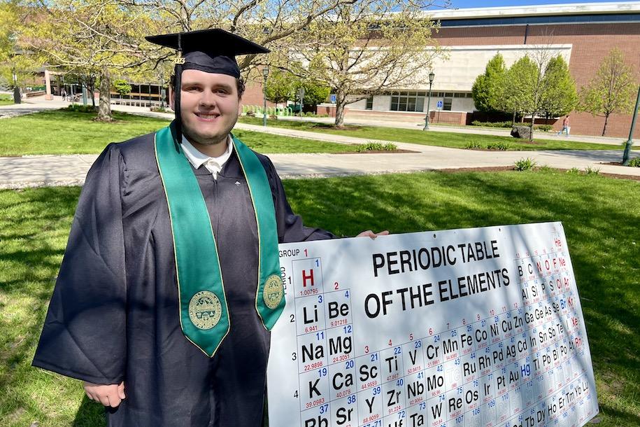 Sam Joyner dressed in UVM regalia holding a large Periodic Table of the Elements 
