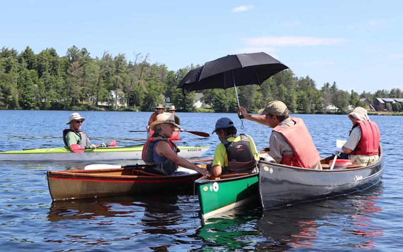Tour participants compare the color of the lake to a water color chart; the shade from an umbrella helps to get an accurate observation. Man in canoe holds up umbrella while another canoeist measures water color on Lower Saint Regis Lake.