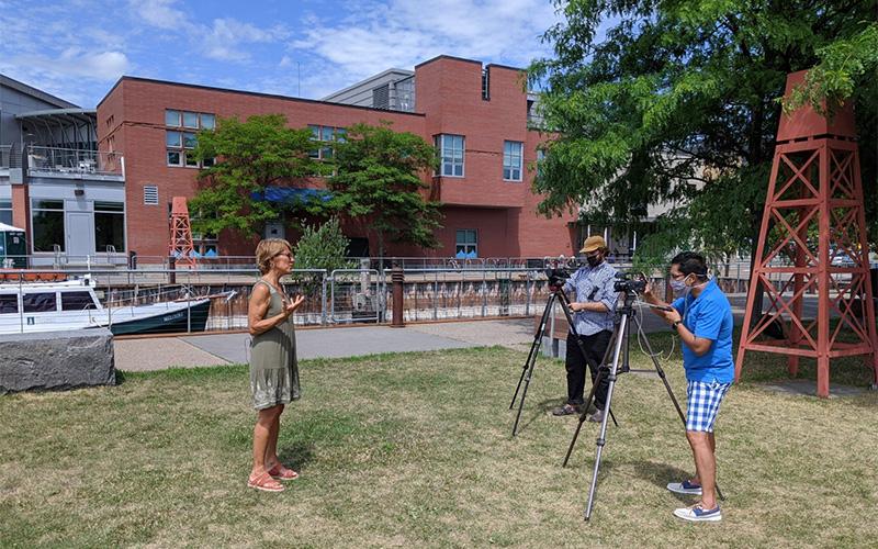 Marco Ayala and Jesse Keefe film Lake Champlain Sea Grant's Linda Patterson. Two videographers film a woman speaking on lawn of building near Lake Champlain