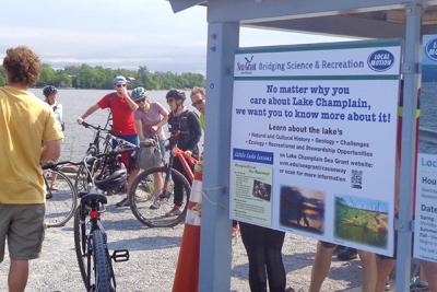 Group of people with bikes on Lake Champlain causeway next to Little Lake Lesson sign