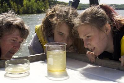 Two students and a teacher in yellow life jackets examine a beaker holding a water sample near a lake