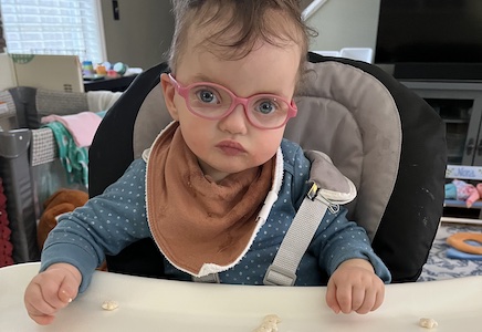 A blue-eyed toddler in pink glasses sits in a high chair in a messy kitchen, looking straight-faced directly at the camera.