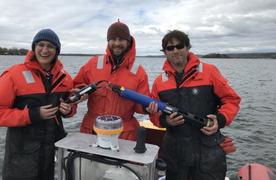 Three UVM researchers on a small boat holding up water quality sensors