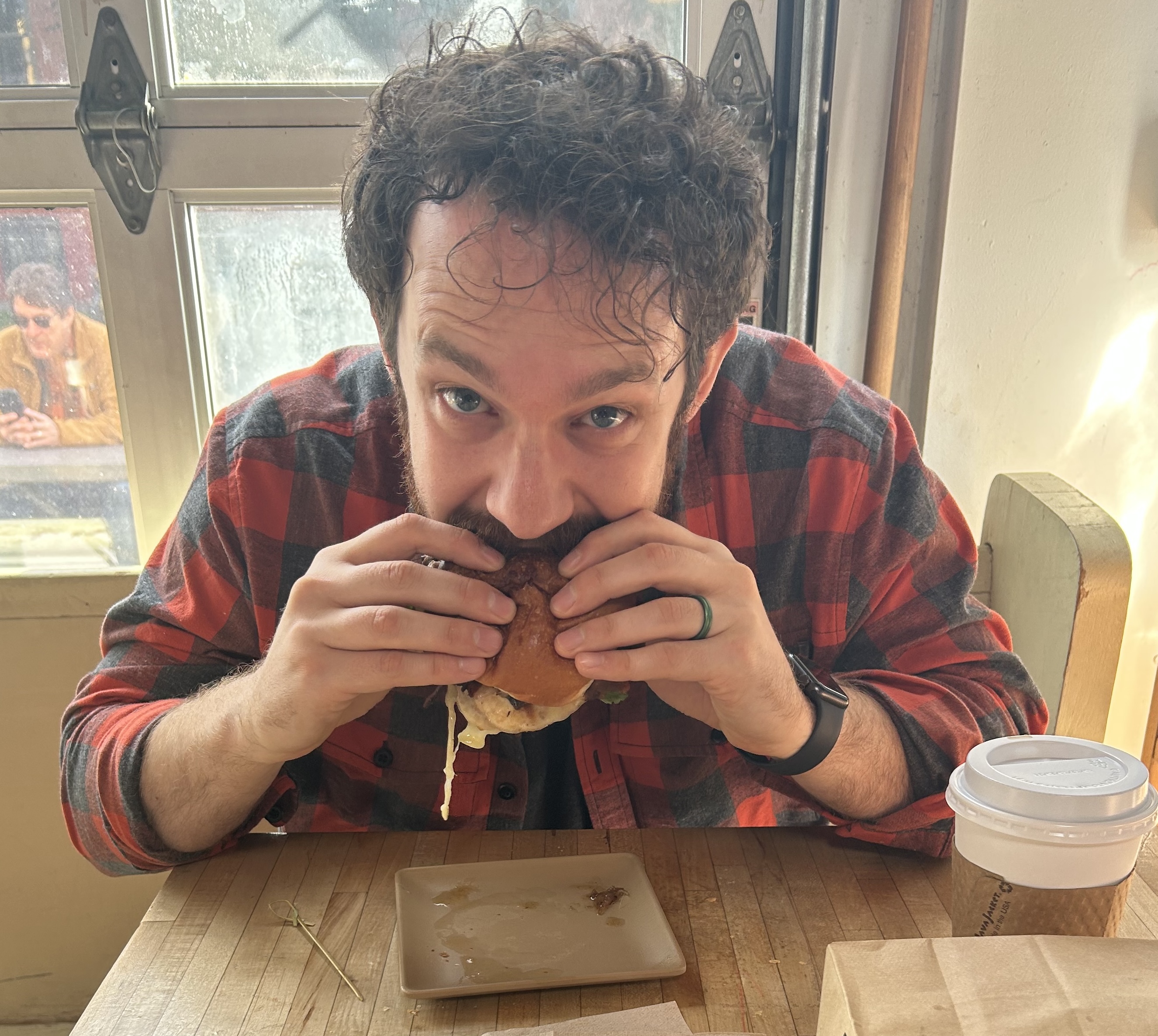 A man in a dark flannel with dark hair eats a breakfast sandwich