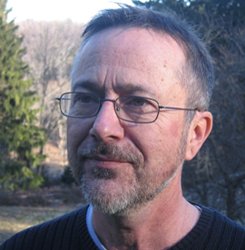 David Lloyd, looking off camera to the left, dark gray hair and salt and pepper short beard, wearing thin dark rimmed glasses and a black shirt