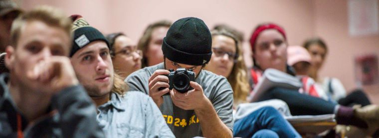 Banner for minor in Reporting and Documentary Storytelling. Student wearing a black beanie sitting at a desk holding a camera pointed at the camera