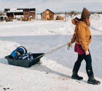 A farmer pulls a baby in a sled with farm outbuildings in the background