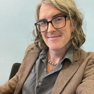 Picture of a white woman with brown hair wearing glasses a gray button up shirt and a brown jacket