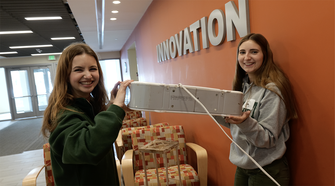 Two students hold a box fan above a tower they have built for competition