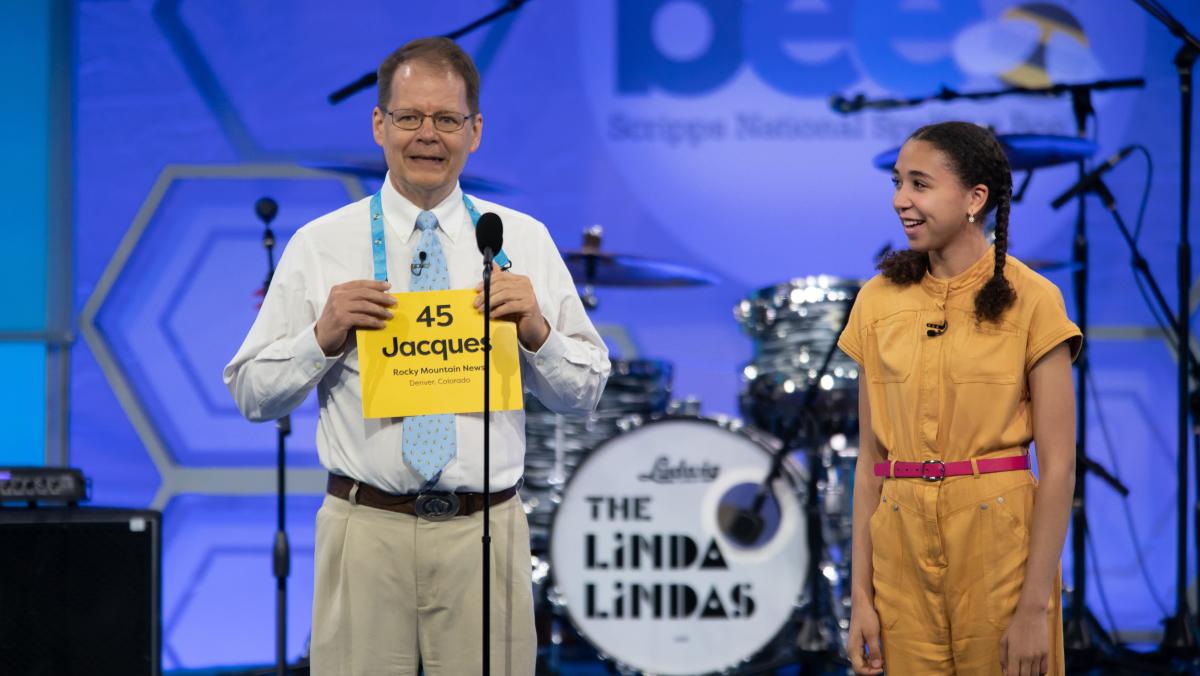 Jacques Bailly stands on stage in front of drum kit at Scripps National Spelling Bee holding sign reading "45 Jacques Rocky Mountain News" Denver Colorado"; he is standing next to a young female Bee contestant