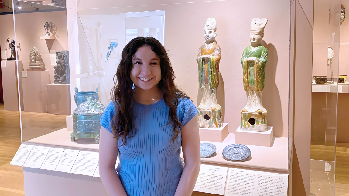 UVM student Zoe Alpert stands in front of a museum case holding two ceramic figures.