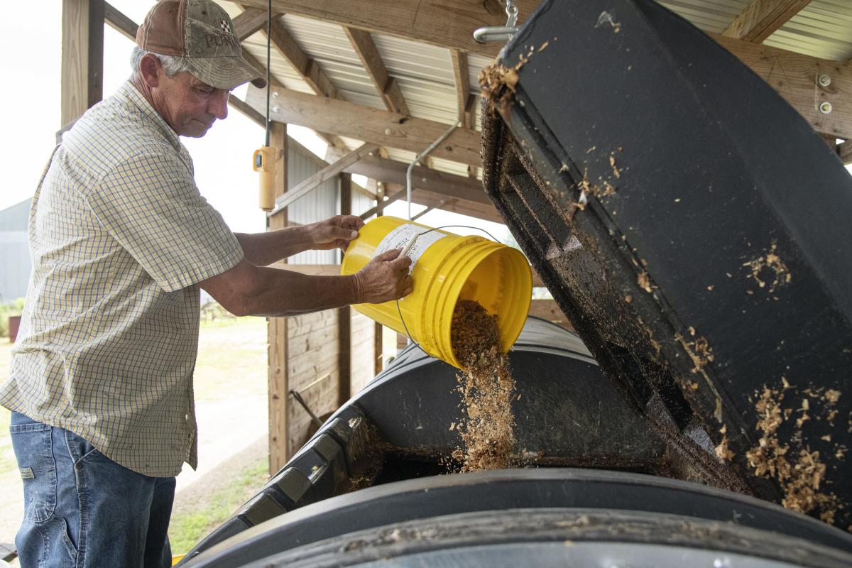 A person dumps leftover food from a bucket into a large composter. 