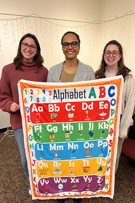 Larner medical students Sofie Leusch ’28, Jasmine Bazinet-Phillips ’25, and Justine Mee, Health Services Coordinator, CVHS, hold up an alphabet blanket provided to families at the Family Health and Literacy Night at Fletcher Free Library this Fall.