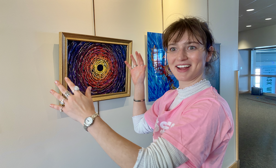 A pale-skinned college aged woman in a pink tshirt and white turtleneck gestures excitedly at a small framed painting of a supernova in reds and yellows on a black background.