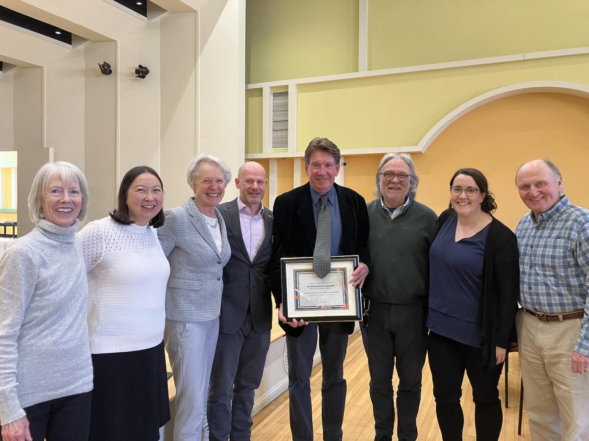 A group of people pose for a photo around a florid man in his mid-sixties holding a framed award