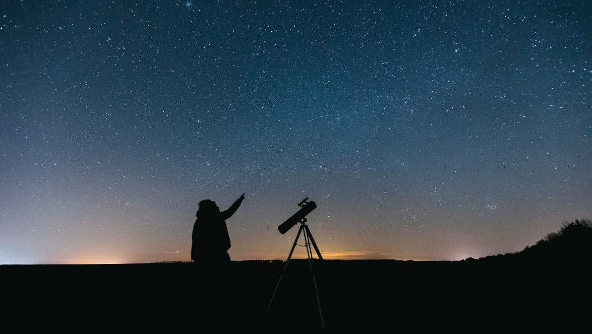 An image of a person beside a telecope. The sky in the background is full of stars, and the person is pointing upward.