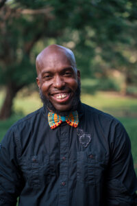 A photo of Bryan Dewsbury smiling to the camera. He is wearying a navy-blue button-up shirt and colorful bowtie