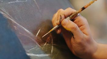 Closeup of a hand holding a paintbrush against a dark canvas.