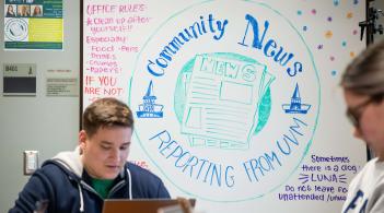 A man focuses on his laptop, sitting in front of a whiteboard. On the board, there is a drawn version of the UVM seal with the words, "Community News: Reporting from UVM."