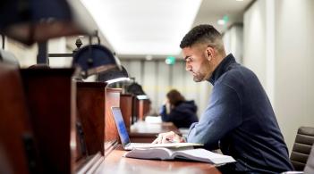 A student studies in the library, hovering over a silver MacBook.
