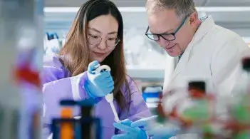 a person in a lab coat oversees a student pipetting liquid into a test tube