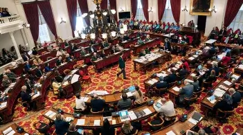 the floor of the state house from above showcasing the many desks, chairs, and people engaged in a legislative session