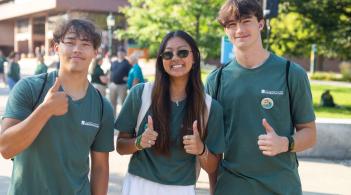 Three students, all wearing the same green Orientation shirt, pose for the camera giving thumbs up