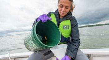 A student sitting on a boat on the lake pouring water from a 5 gallon bucket into a funnel