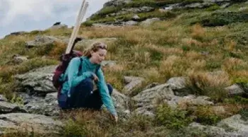 A student crouching on a mountainside looking at plant growth, she is wearing a backpack with equipment in it.