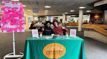 Tutors Symaira and Max hosted our annual International Writing Centers Week table on February 14th. They sit behind a table draped with a green tablecloth with the writing centers logo with students sitting at library tables behind them.