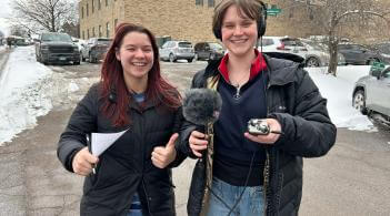 Two university of Vermont students wearing coats hold a microphone and notes while they cover the 2024 federal election.