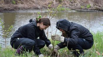 Two students planting a tree in front of a river flowing in the background.