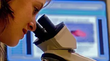 Individual looking through a microscope in front of a background of an image on a computer screen