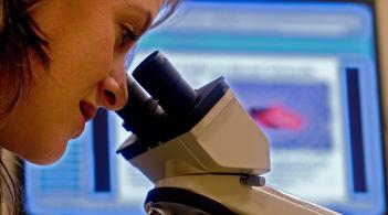 Individual looking through a microscope in front of a background of an image on a computer screen