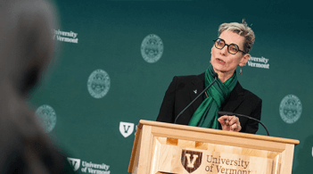 woman speaks at a wooden podium in front of a green step and repeat