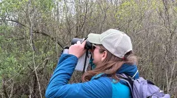 A woman in a blue jacket and hat looks through binoculars at trees and shrubs.