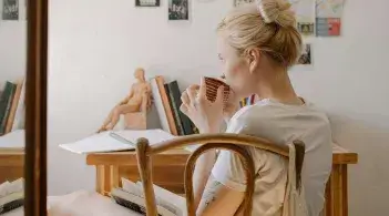 A blonde white college student sits in a chair at her dorm desk, contemplating something off camera