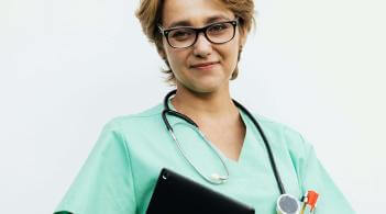 A Latina woman with blonde hair and glasses, wearing green medical scrubs, holds a clipboard