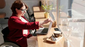 A white woman in her twenties with glasses and curly chestnut hair sits in a wheelchair, setting up a laptop at a café.