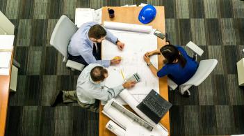 Three people sit around a table in an office, looking at a blueprint.