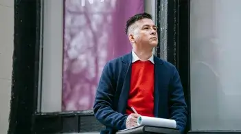 A Latino man in a red sweater and sport jacket stops to ponder while taking notes on a pad in his lap