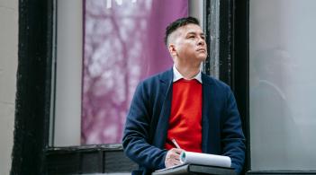 A Latino man in a red sweater and sport jacket stops to ponder while taking notes on a pad in his lap