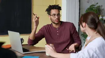 A copper-skinned man with glasses and short dreadlocs explains something to a white woman sitting next to him