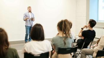 A white man lectures to a group of college-aged students in folding chairs.