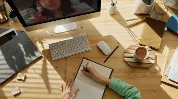 A student sits taking notes in front of a computer screen, showing another student