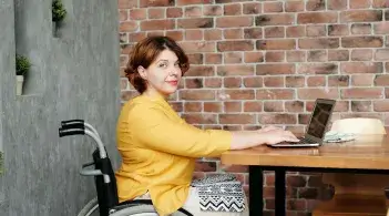 A pale-skinned woman in a wheelchair sits at a table, hands on an open laptop, and looks knowingly at the camera