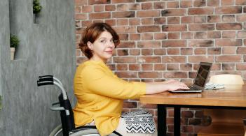 A pale-skinned woman in a wheelchair sits at a table, hands on an open laptop, and looks knowingly at the camera