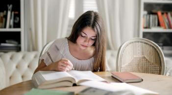 A pale-skinned college-aged girl writes in a notebook at a table filled with books